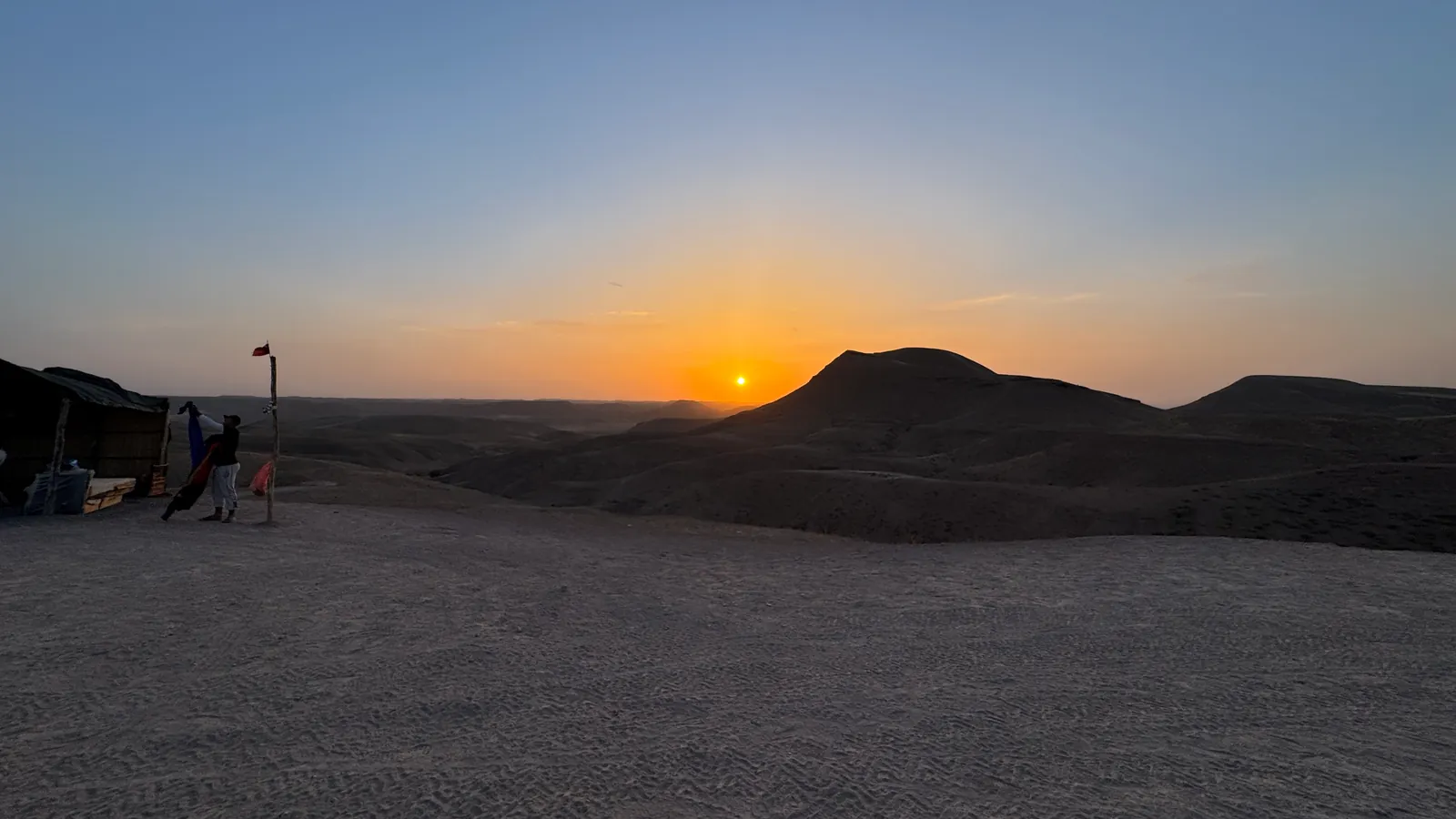 Le soleil bas sur l'horizon, à côté d'une colline sombre du désert d'Agafay ; à gauche, une tente, un mât à fanion rouge et une silhouette