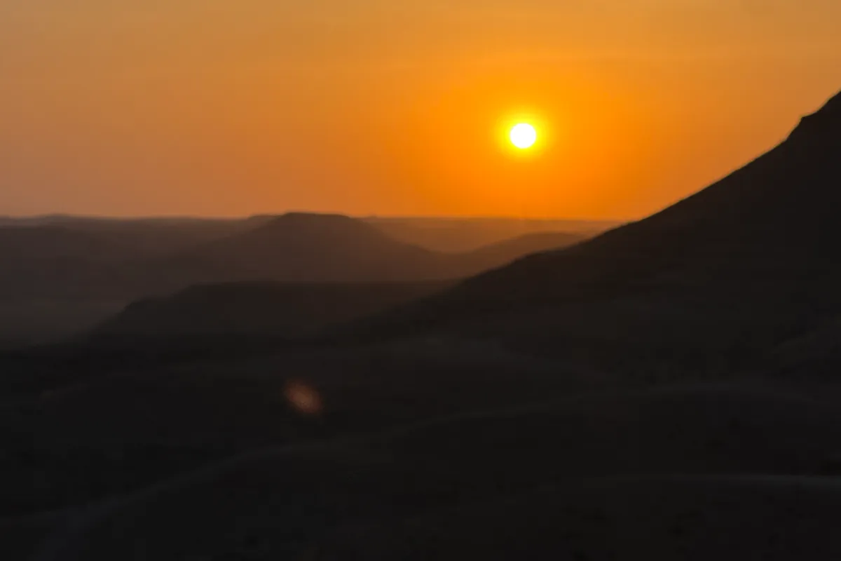 The orange sun above the already dark hills of the Agafay desert, with a shaded slope in the foreground (June 2026)