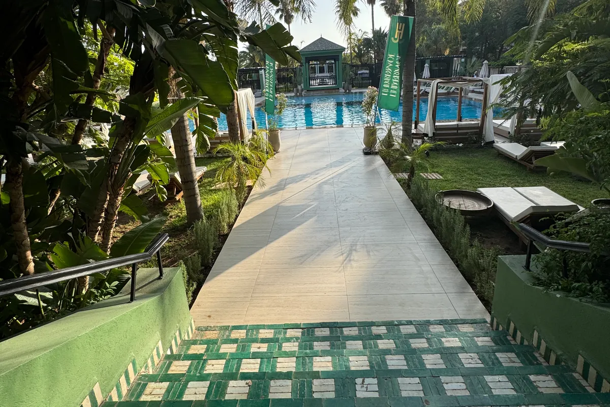 Des marches en zellige vert et blanc ouvrent sur une allée dallée qui mène à la piscine, entre palmiers et plantes à larges feuilles (juin 2026)