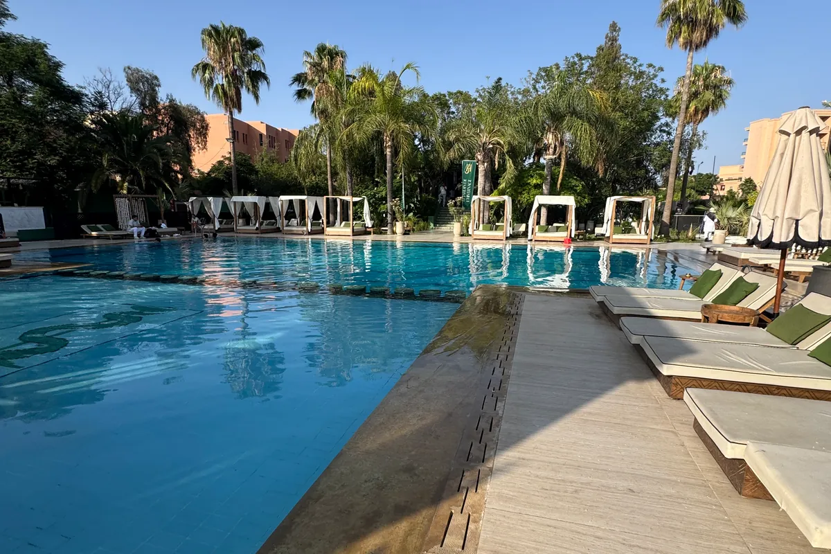 Le bord de la grande piscine, avec des transats à coussins verts, des lits à baldaquin blancs et des palmiers, au Jardin de l'Hivernage (juin 2026)