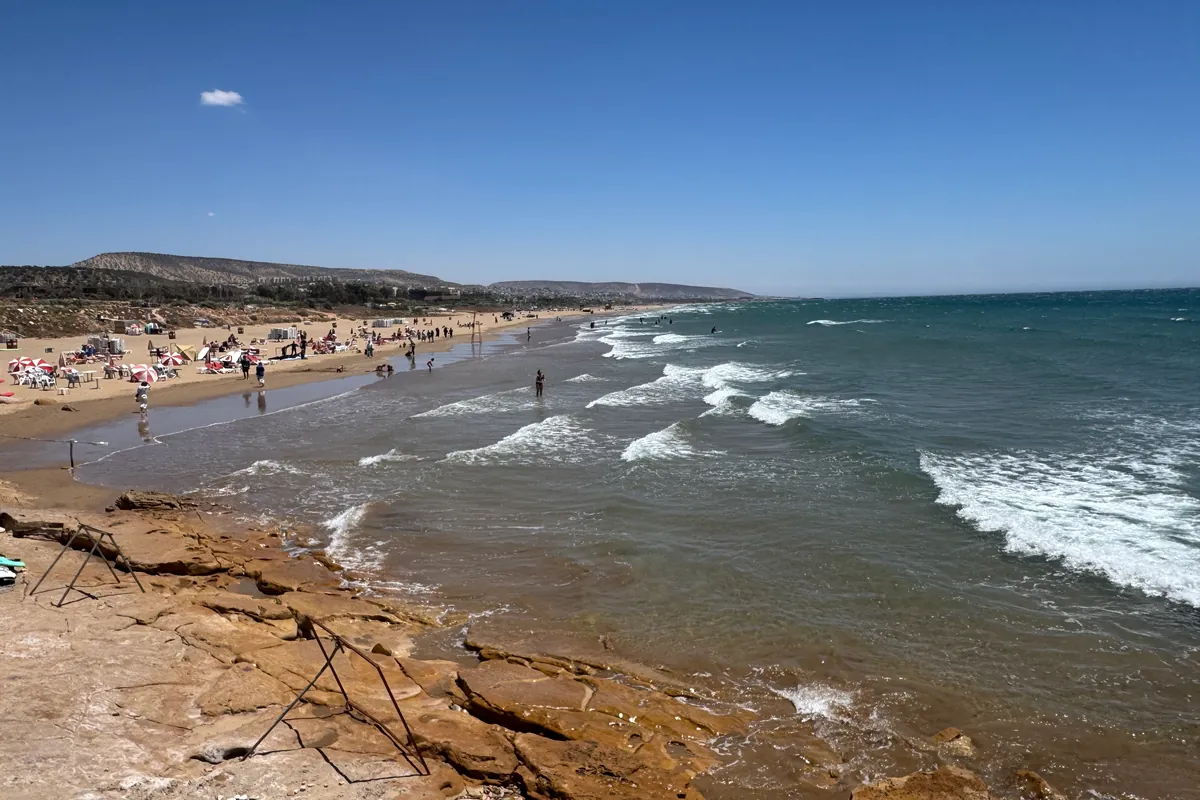 Las rocas al borde de la gran playa de Taghazout, con sus olas y sus bañistas, mirando hacia Taghazout Bay (mayo de 2026)