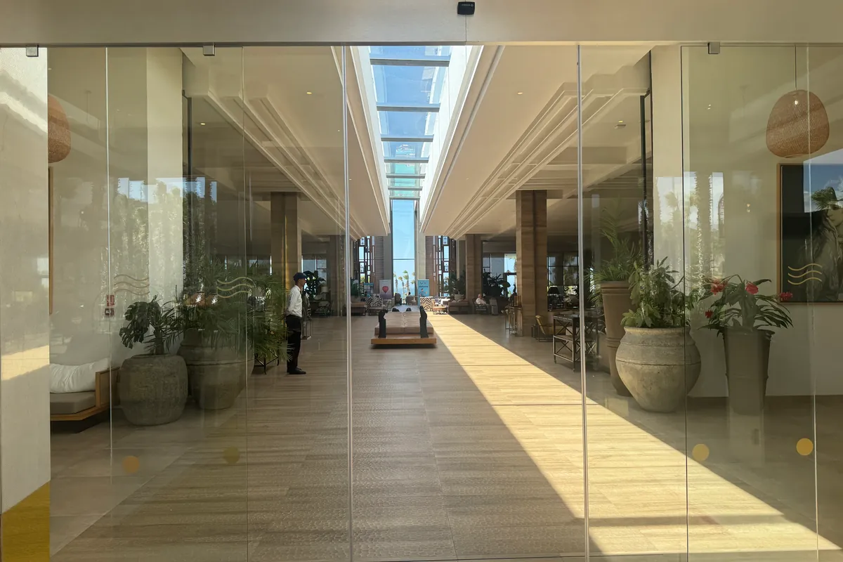 El vestíbulo del hotel tras unas puertas de cristal: un largo pasillo bajo una cubierta de cristal, con grandes tinajas y plantas en macetas (mayo de 2026)