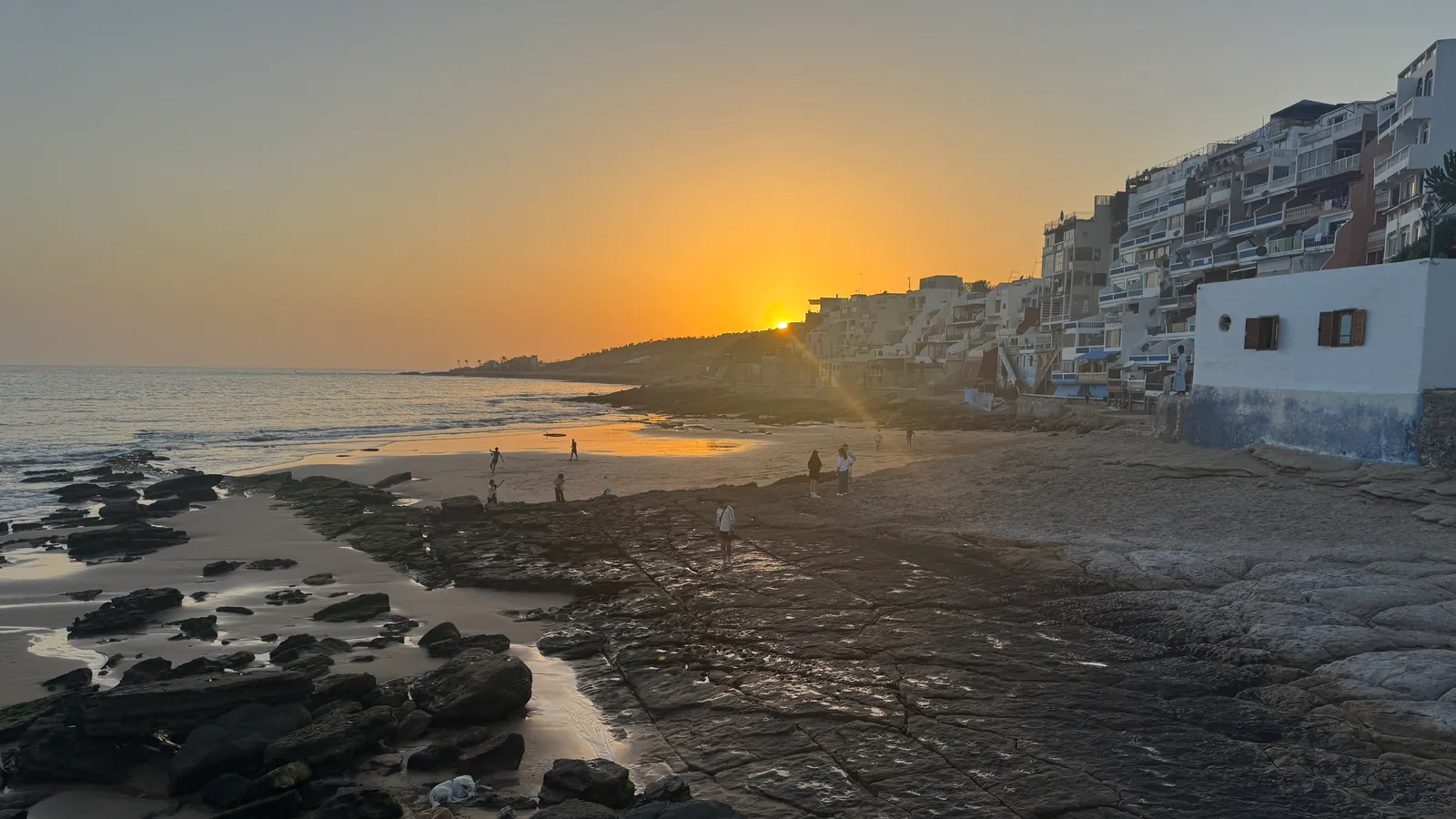 Le soleil descend derrière la colline au bout de la plage ; au premier plan, les rochers et la dalle mouillée de Taghazout, au pied des immeubles du front de mer