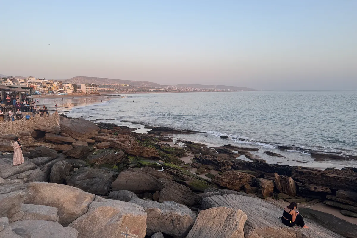 Une terrasse sous paillotes, pleine de monde, sur les rochers de Taghazout ; au-delà, la plage, les immeubles du village et la baie (mai 2026)