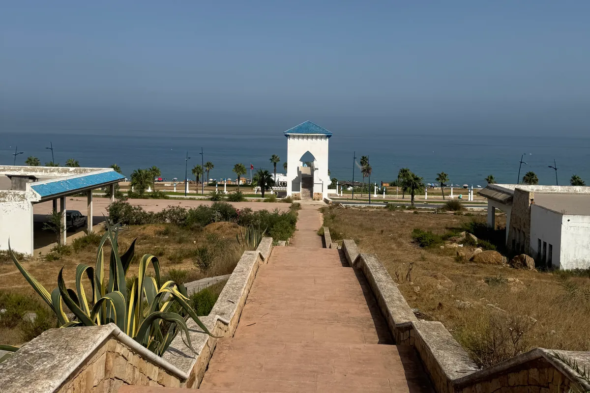 Un long escalier bordé de murets de pierre descend, près d'un grand agave, vers la tour blanche d'une passerelle ; derrière, la route côtière, la plage et la mer (septembre 2026)