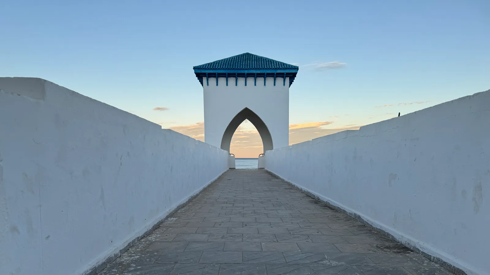 Entre deux parapets blancs, le tablier dallé d'une passerelle mène à une tour coiffée de tuiles bleu-vert, dont l'arche en ogive s'ouvre sur la mer en fin de journée