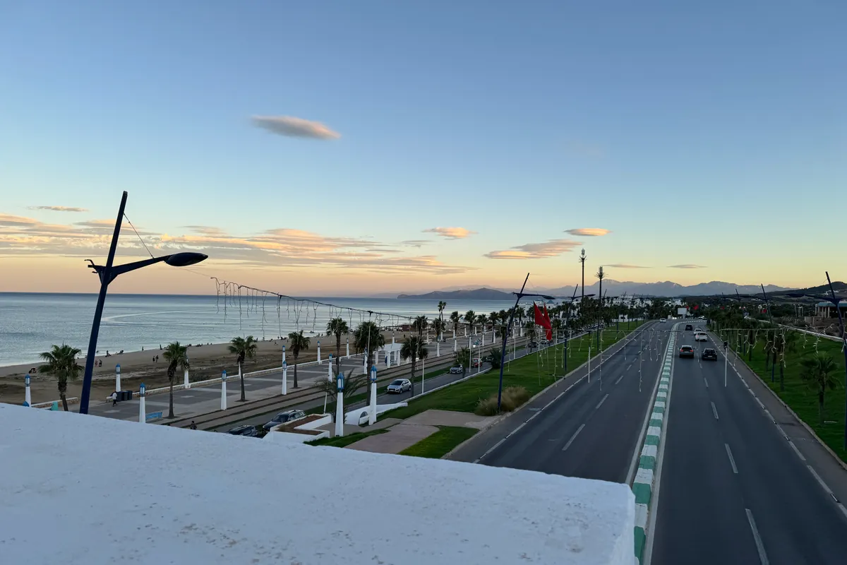 Depuis la passerelle : la plage Riffiine, sa promenade bordée de palmiers et de lanternes, la route côtière et les montagnes au loin, en fin de journée (septembre 2026)