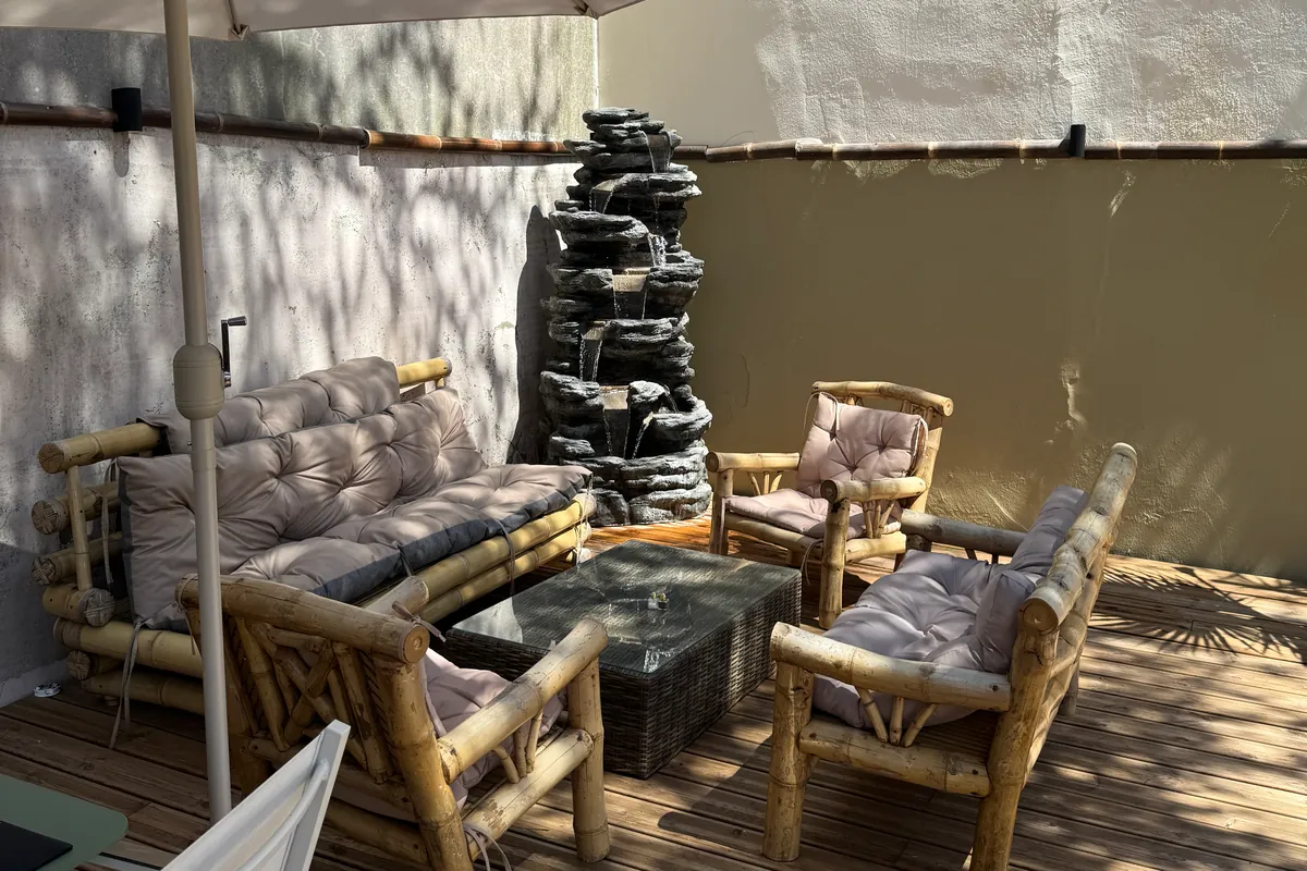 Un coin salon de la terrasse du Z : un canapé et des fauteuils en bambou à coussins, une table basse et une petite fontaine en cascade imitant la roche (juillet 2026)