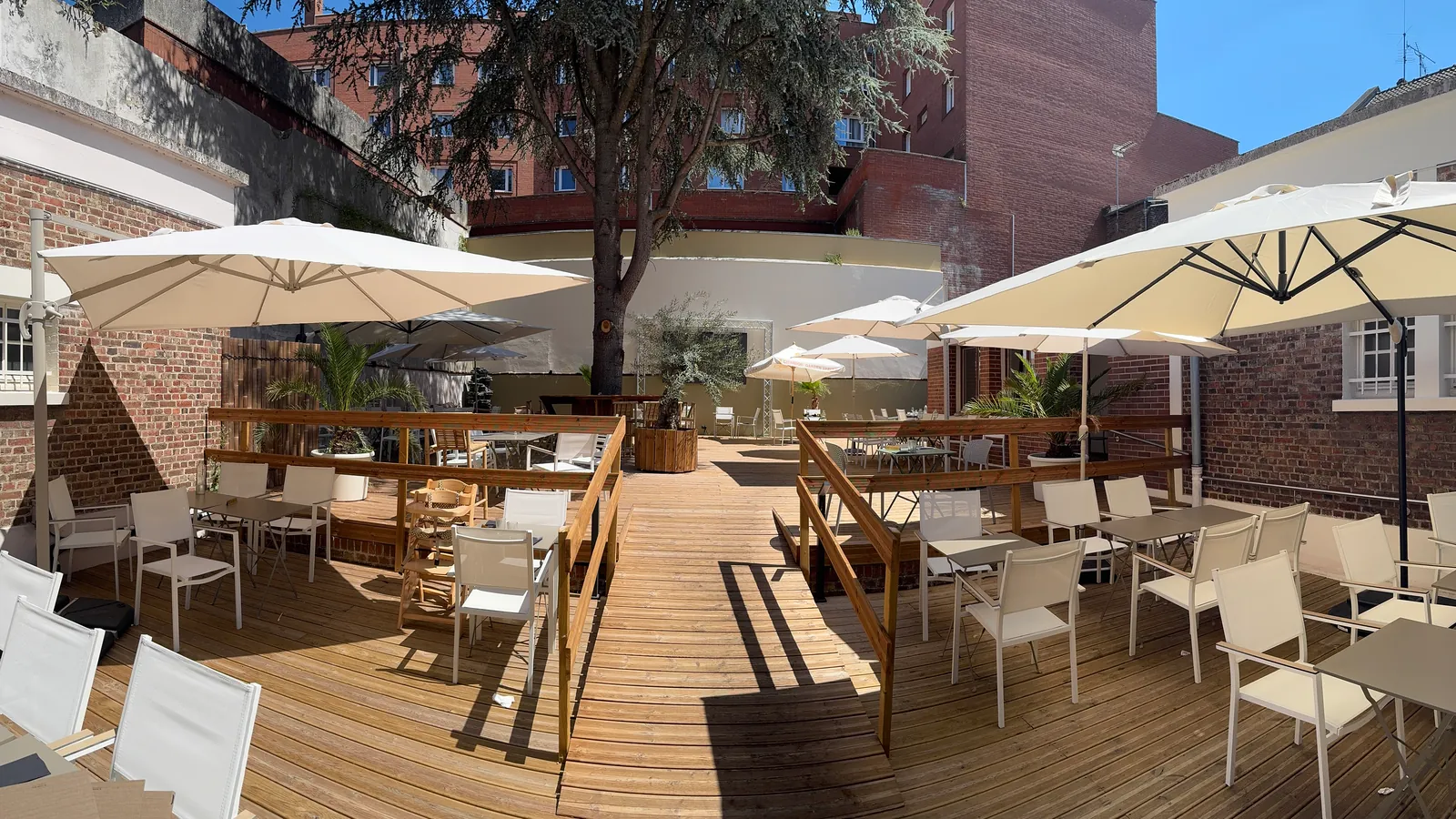 La terrasse du Z en plein jour : un plancher de bois, des tables grises, des chaises blanches, de grands parasols et un grand arbre