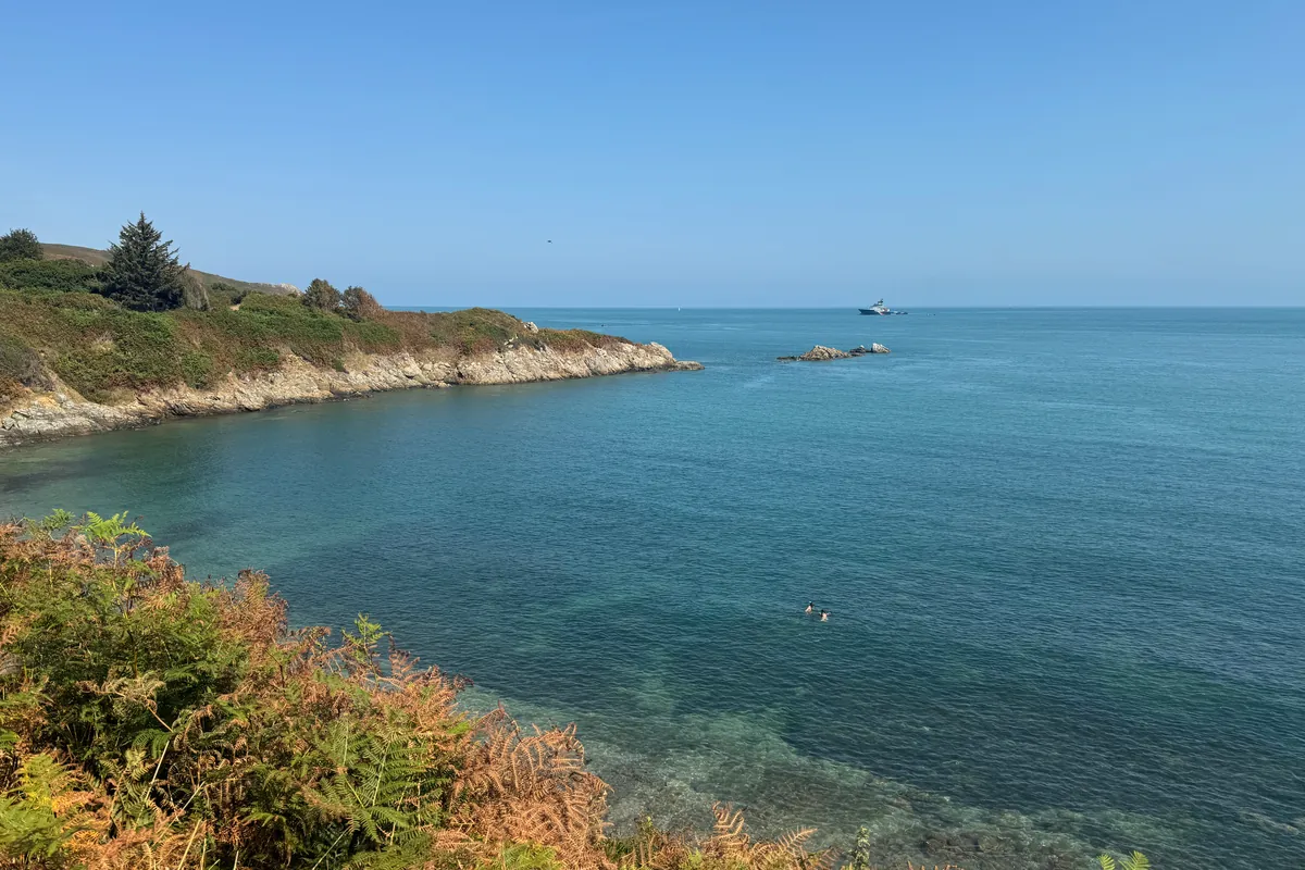 L'eau turquoise de la baie de Quervière, entre une pointe de lande et des rochers, avec deux nageurs au loin (août 2026)