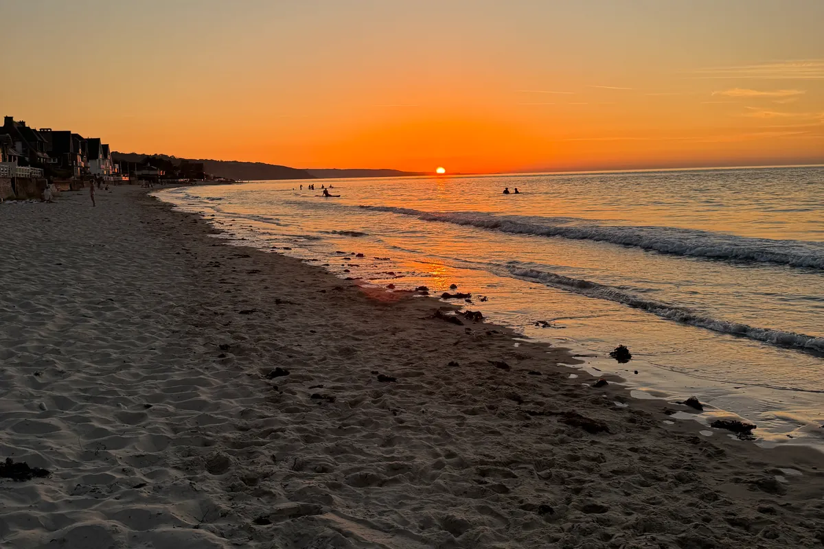 Le soleil se pose sur l'horizon, devant des baigneurs en silhouette, sur la plage d'Urville-Nacqueville (août 2026)
