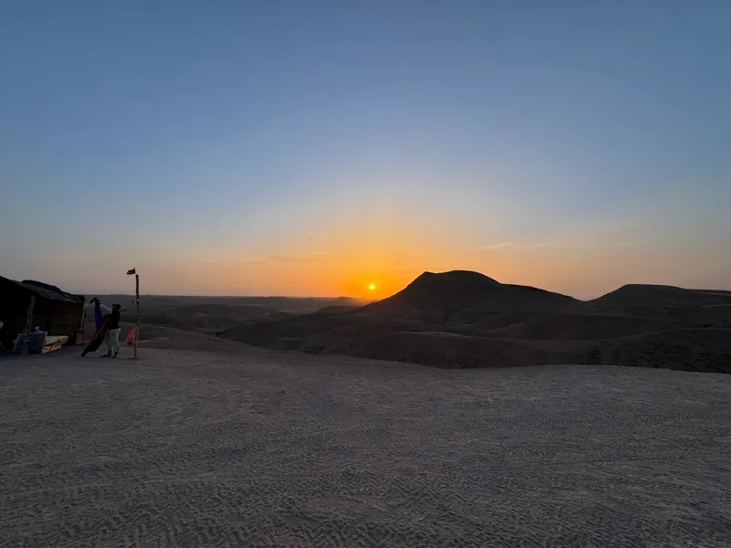 Le soleil bas sur l'horizon, à côté d'une colline sombre du désert d'Agafay ; à gauche, une tente, un mât à fanion rouge et une silhouette