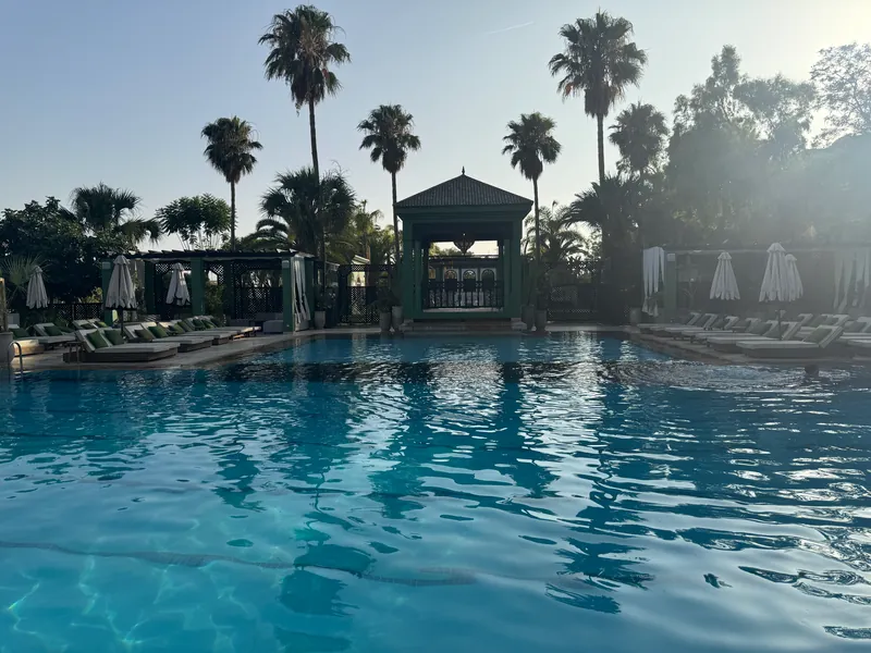 La piscine du Jardin de l'Hivernage face à un pavillon vert à toit pyramidal, entre palmiers, transats à coussins verts et parasols fermés