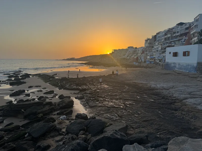 Le soleil descend derrière la colline au bout de la plage ; au premier plan, les rochers et la dalle mouillée de Taghazout, au pied des immeubles du front de mer