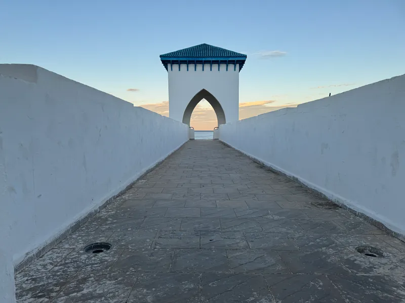 Entre deux parapets blancs, le tablier dallé d'une passerelle mène à une tour coiffée de tuiles bleu-vert, dont l'arche en ogive s'ouvre sur la mer en fin de journée