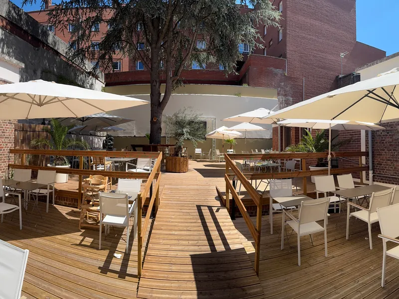 La terrasse du Z en plein jour : un plancher de bois, des tables grises, des chaises blanches, de grands parasols et un grand arbre